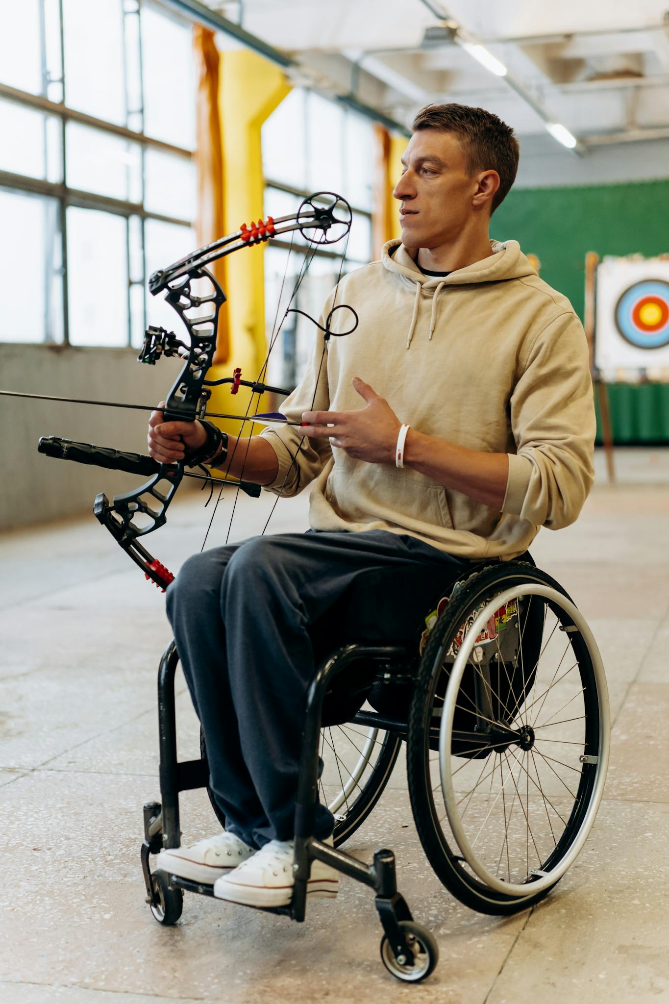 A male archer in a wheelchair practicing archery indoors, aiming his bow at a target.