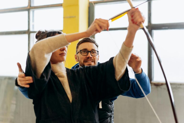 Adult woman learning archery with guidance from male coach indoors.