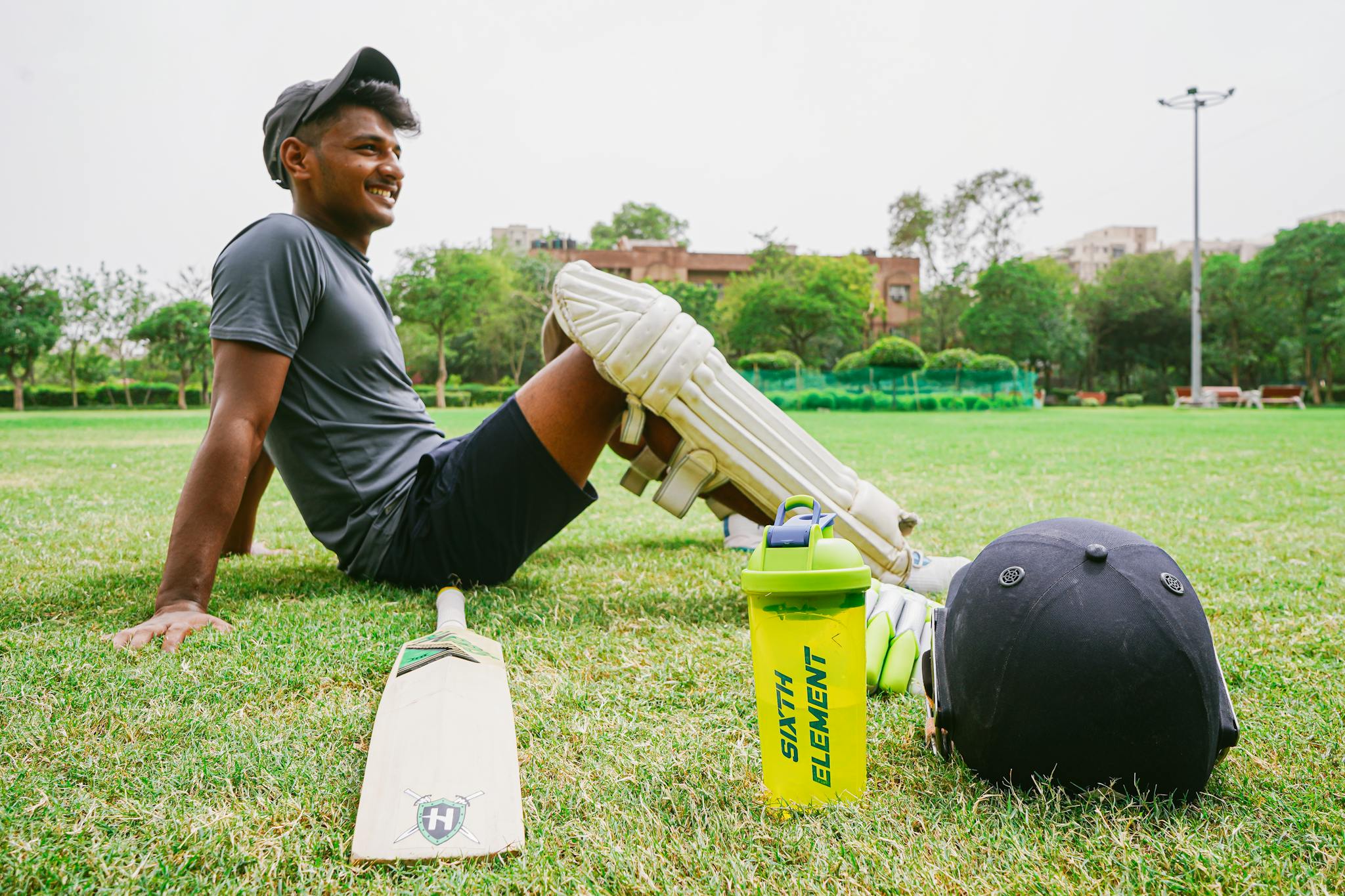 Side view of full body happy sportsman wearing sportswear and cricket pads resting on lawn beside cricket bat and helmet and sport water bottle